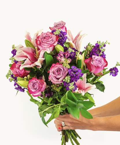 A vibrant bouquet featuring pink lilies, purple flowers, and green foliage, elegantly held.