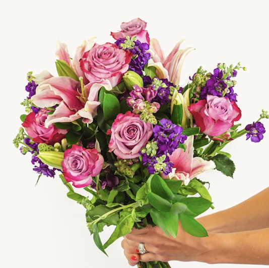 A vibrant bouquet featuring pink lilies, purple flowers, and green foliage, elegantly held.
