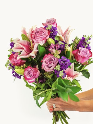 A vibrant bouquet featuring pink lilies, purple flowers, and green foliage, elegantly held.