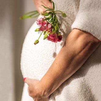 Gentle hand on a pregnant belly, holding delicate pink flowers in a cozy setting.