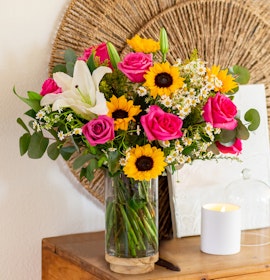 Vibrant floral arrangement featuring pink roses, sunflowers, and white lilies in a glass vase.