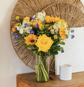 Bright bouquet of yellow roses, sunflowers, and blue flowers in a stylish glass vase.