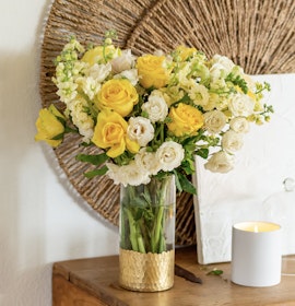 A vibrant bouquet of yellow roses and white flowers in a glass vase, complemented by a gold base.