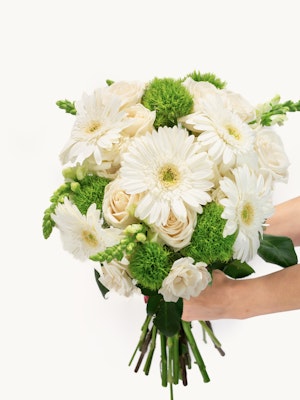 Delicate white gerbera daisies and soft roses in a lush green bouquet held gracefully.