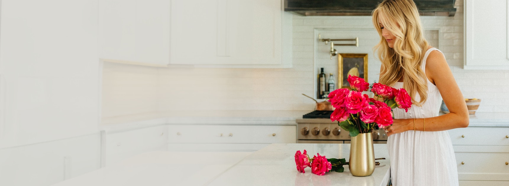 A woman in a white dress arranges vibrant pink roses in a gold vase on a kitchen counter.