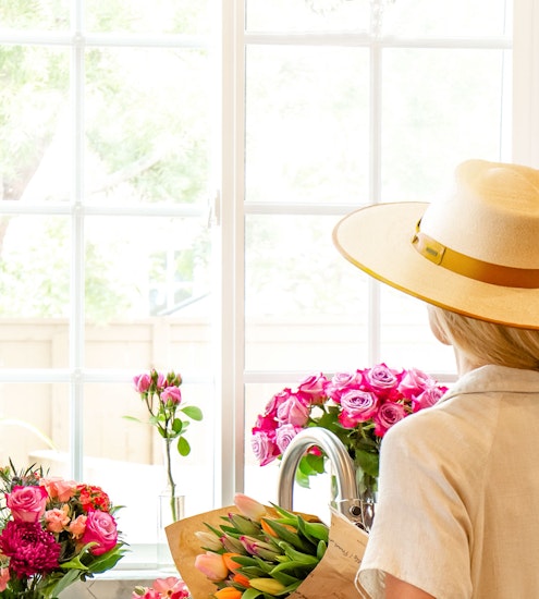 A woman in a sun hat admires vibrant flowers by a bright window, creating a cheerful ambiance.